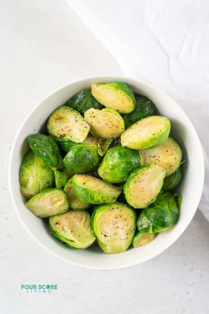 photo of a bowl of steamed brussels sprouts with salt and pepper on top, with a white kitchen towel next to the bowl