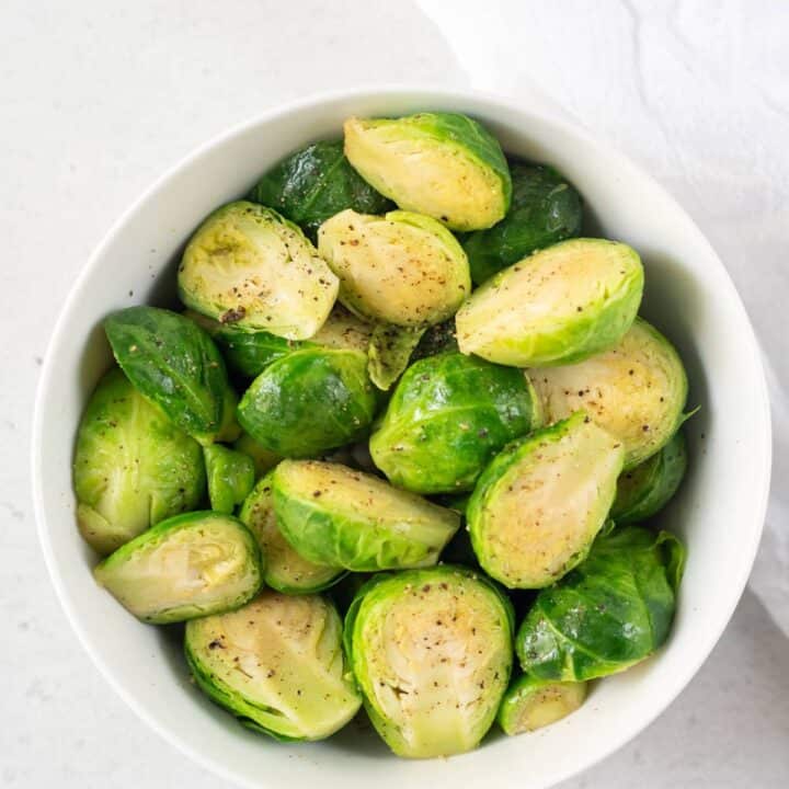 photo of a bowl of steamed brussels sprouts with salt and pepper on top, with a white kitchen towel next to the bowl