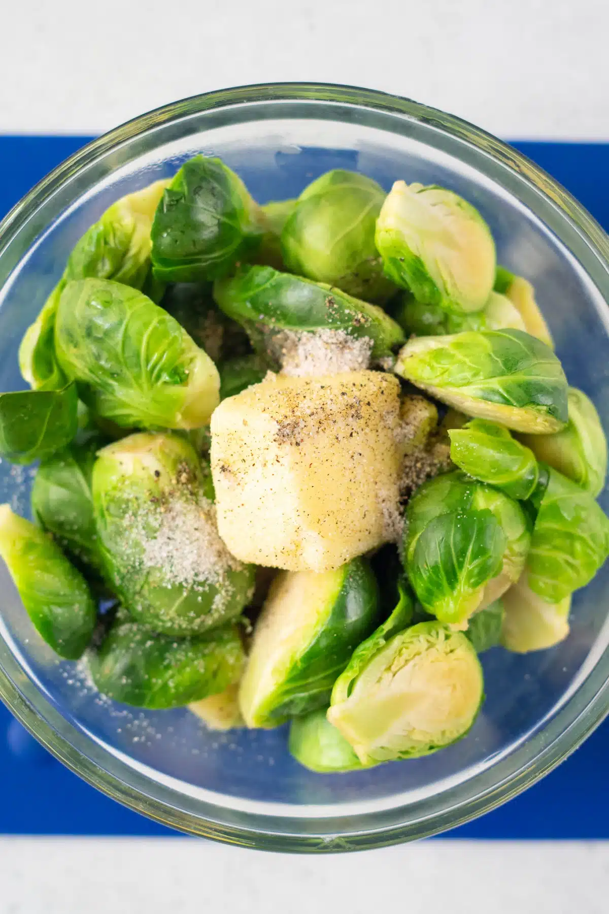 closeup photo of steamed brussels sprouts in a clear glass bowl, with butter, salt, and pepper on top, ready to be mixed together