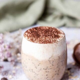 photo of tiramisu overnight oats in a glass mug surrounded by decorative flowers and a green cloth in the background