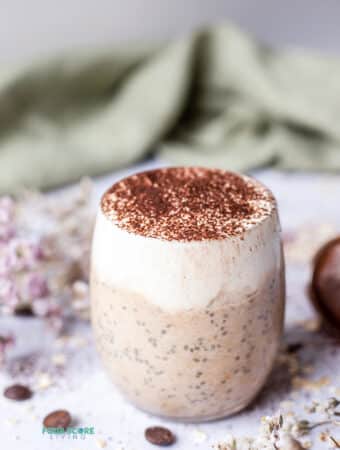 photo of tiramisu overnight oats in a glass mug surrounded by decorative flowers and a green cloth in the background