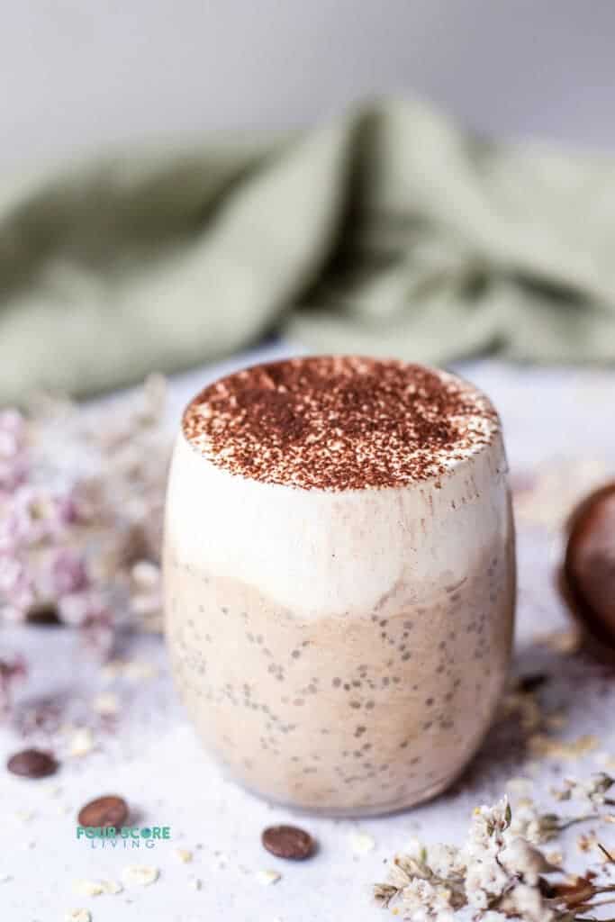 photo of tiramisu overnight oats in a glass mug surrounded by decorative flowers and a green cloth in the background