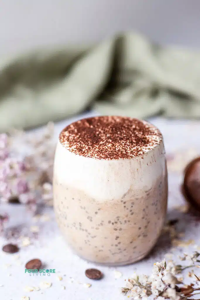 photo of tiramisu overnight oats in a glass mug surrounded by decorative flowers and a green cloth in the background