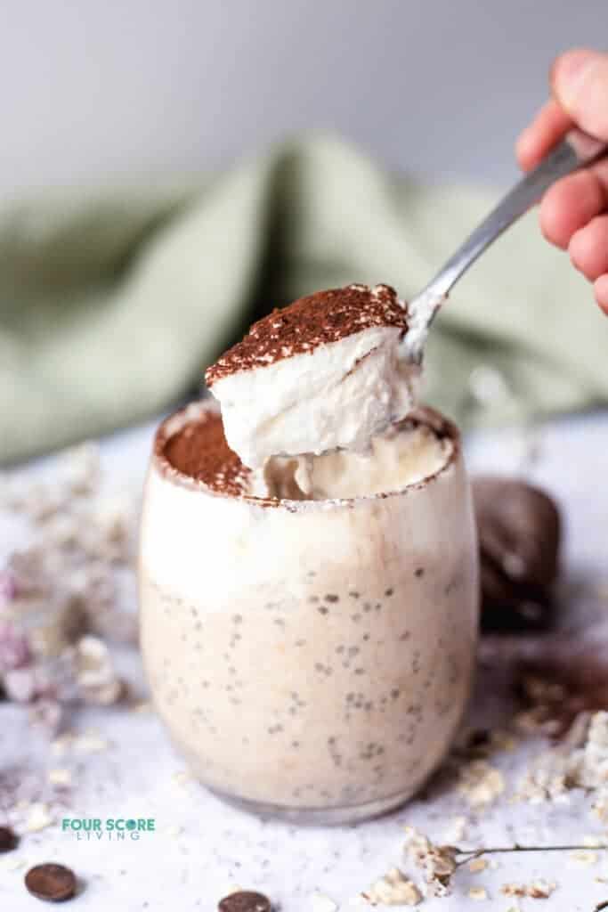 photo of a hand using a spoon to scoop tiramisu overnight oats out of a glass mug, with a green kitchen towel in the background