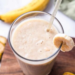 Top view photo of a glass of Banana Juice with a straw. The glass is on a wooden cutting board surrounded by slices of banana.