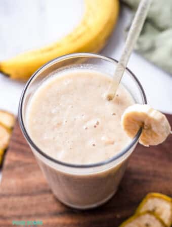 Top view photo of a glass of Banana Juice with a straw. The glass is on a wooden cutting board surrounded by slices of banana.