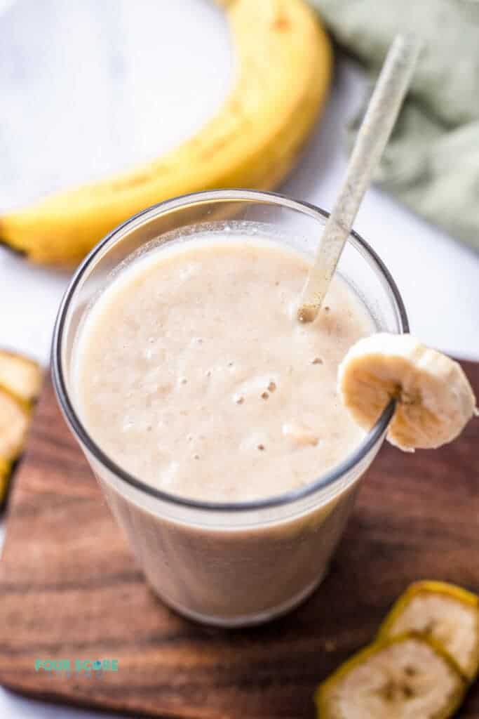 Top view photo of a glass of Banana Juice with a straw. The glass is on a wooden cutting board surrounded by slices of banana. 