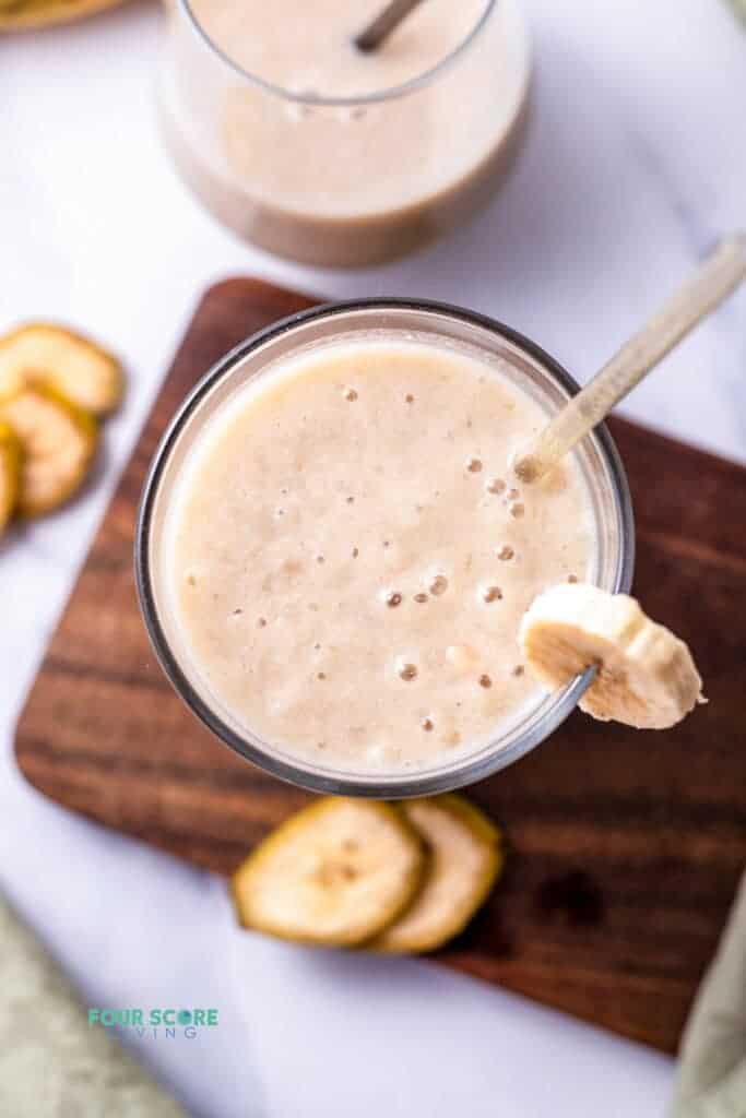 Top view photo of a glass of Banana Juice with a straw. The glass is on a wooden cutting board surrounded by slices of banana.