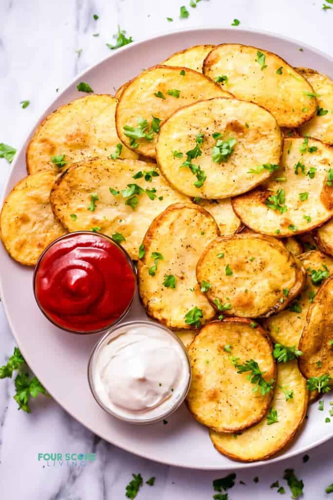 Top view photo of Cottage Fries on a white plate, with 2 sides of dipping sauces. The fries are garnished with chopped fresh parsley. 