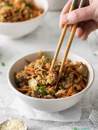 Photo of a hand holding a pair of chopsticks, and picking up a bite of Egg Roll in a Bowl from a white bowl. There is another white bowl in the background.