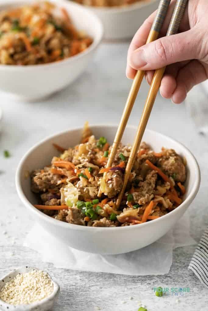 Photo of a hand holding a pair of chopsticks, and picking up a bite of Egg Roll in a Bowl from a white bowl. There is another white bowl in the background. 