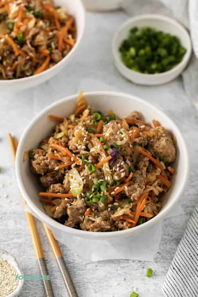 Photo of a white bowl filled with this Egg Roll in a Bowl recipe. There are a pair of chopsticks next to the bowl and a small bowl with sliced green onions in the background.