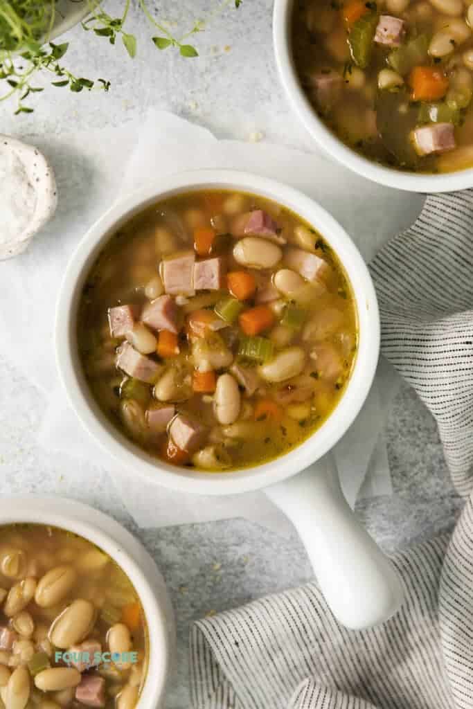 Top view photo of 3 bowls of Ham and White Bean Soup, ready to eat. There is a striped kitchen towel by the bowls. 