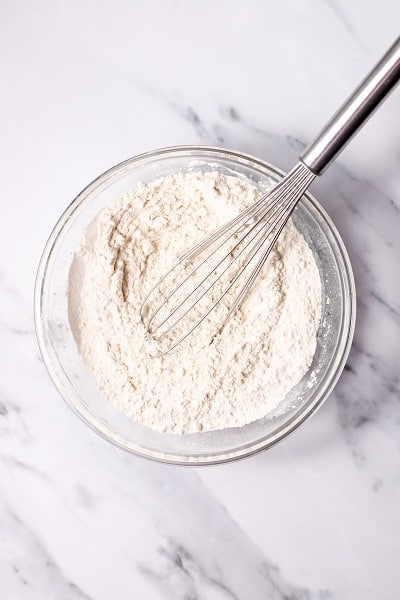Top view photo of a clear bowl with flour, baking soda, and salt mixed together with a wire whisk.