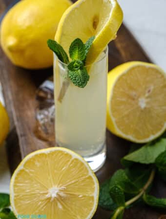 Photo of a White Tea Shot in a wooden cutting board. There is a slice of lemon and sprig of mint in the shot, and the glass is surrounded by mint, lemons, and ice.