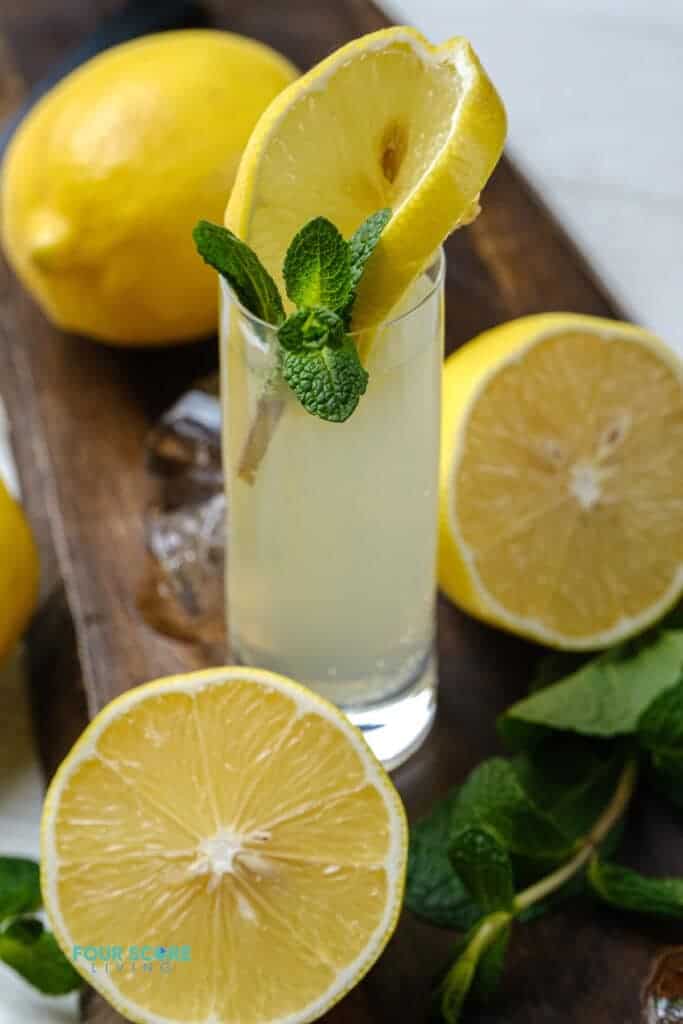 Photo of a White Tea Shot in a wooden cutting board. There is a slice of lemon and sprig of mint in the shot, and the glass is surrounded by mint, lemons, and ice. 