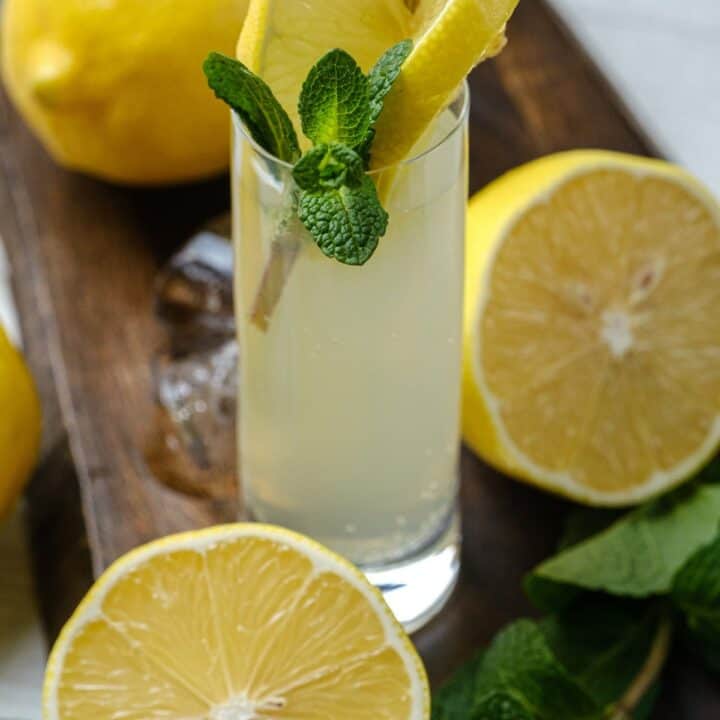 Photo of a White Tea Shot in a wooden cutting board. There is a slice of lemon and sprig of mint in the shot, and the glass is surrounded by mint, lemons, and ice.