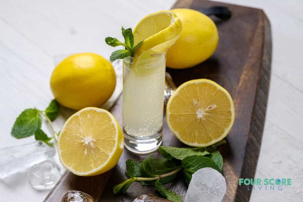 Photo of a White Tea Shot in a wooden cutting board. There is a slice of lemon and sprig of mint in the shot, and the glass is surrounded by mint, lemons, and ice.