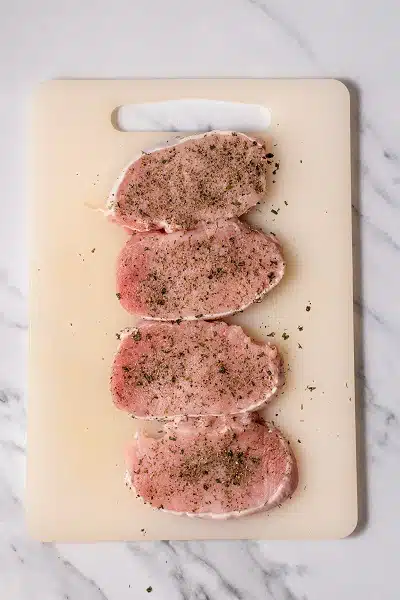 Top view photo of a cutting board with 4 pork chops, and seasoning sprinkled over each pork chop.