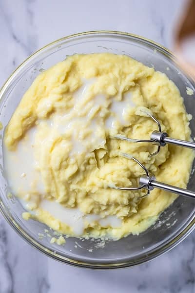 Top view photo of a clear bowl filled with boiled potatoes and milk, and being mixed by a handheld mixer until creamy.