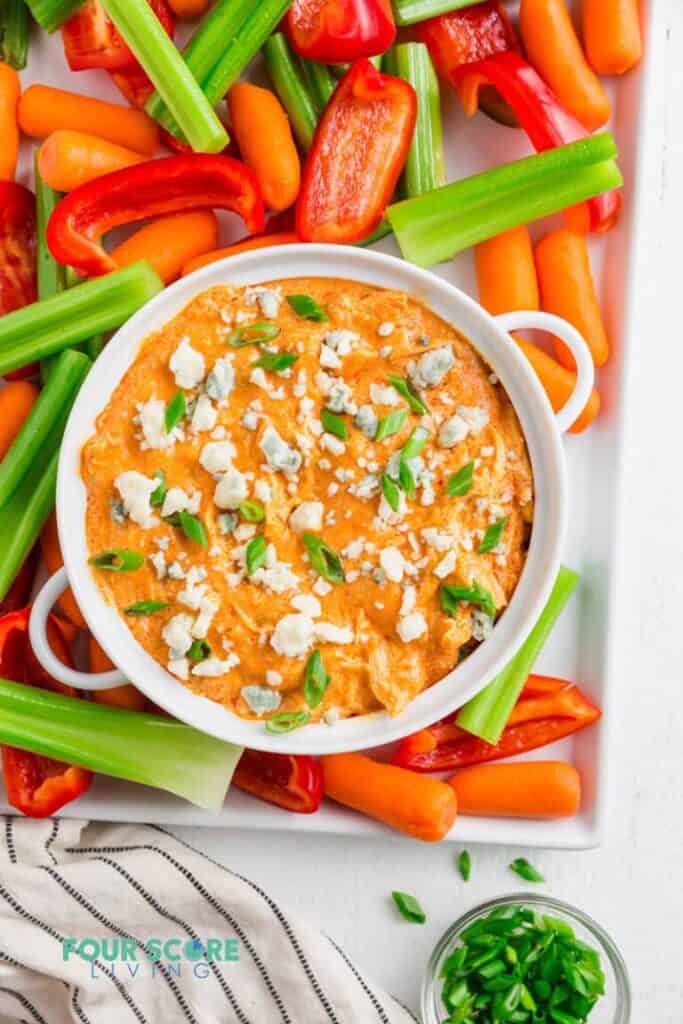 Keto-Buffalo-Chicken-Dip-1.2-768x1152 in a white round dish with handles surrounded by raw vegetable sticks including celery, carrots and peppers. 