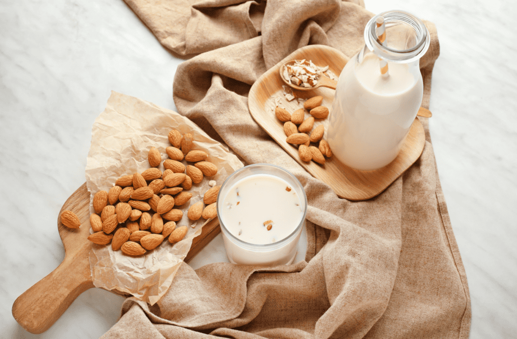 glass jar of almond milk with a straw on a brown plate with whole almonds and a wooden spoon with chopped almonds. There is also a wooden cutting board with whole almonds and a glass of almond milk.