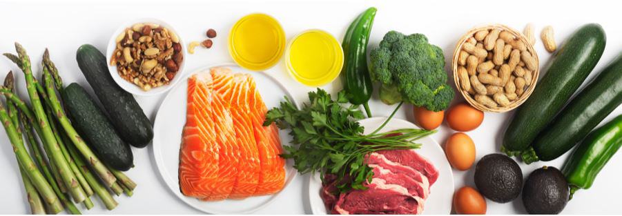keto vegetables such as asparagus, cucumbers, jalapeno, broccoli, zucchini, avocado, parsley spread on a white counter top. There is also a bowl of assorted keto nuts plus keto salmon filets on a white plate and slices of raw keto beef on a white plate. 2 glass bowls full of oil and 4 eggs