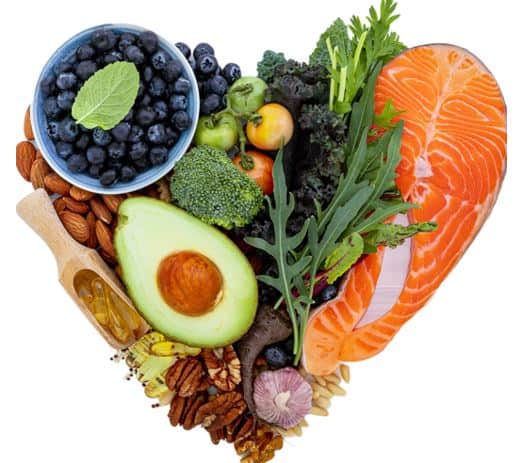 Keto Foods arranged in the shape of a heart, foods include raw salmon steak, fresh baby greens of chard and dandelion, broccoli, a whole purple garlic bulb, a halved avocado with the seed intact, whole almonds, pecans and pine nuts, a small dish of fresh blueberries, a wooden scoop holding yellow gel oil pills, and tiny apples.   