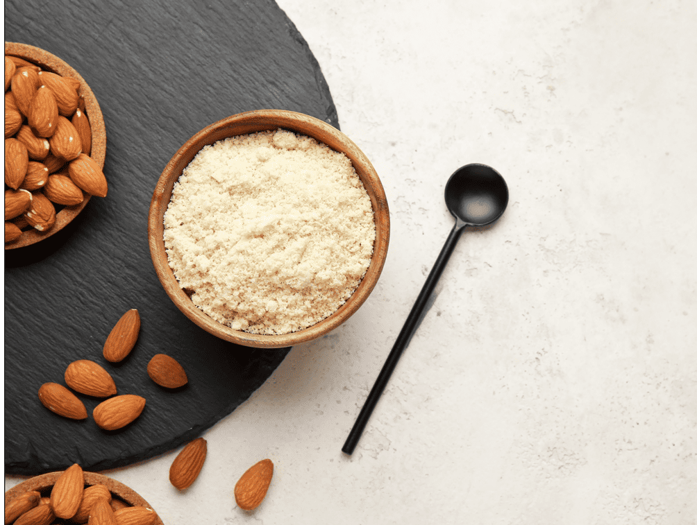 Arial view: medium brown wooden dish filled with ground almond flour and a medium bowl of whole almonds with several spilled onto the surface on a round slate platter resting on an ivory stone surface with a long handled and long bowed dark grey spoon resting on the ivory stone surface. 