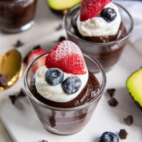 Glass dessert cups of chocolate avocado pudding garnished with whipped cream, 2 blueberries and a halved strawberry. The glass dessert cups are placed on a white square platter with halved fresh Hass avocados, a brass spoon and scattered chocolate chips in the background.