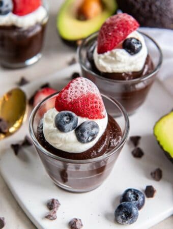 White square plate with 2 glass dessert cups filled with chocolate avocado pudding. The pudding is topped with a dollop of whipped cream, a half of a strawberry and two blueberries. In the background fresh, halved Hass avocados are pictured as well a scattered chocolate chips and a brass spoon.