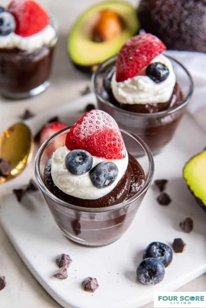 White square plate with 2 glass dessert cups filled with chocolate avocado pudding. The pudding is topped with a dollop of whipped cream, a half of a strawberry and two blueberries. In the background fresh, halved Hass avocados are pictured as well a scattered chocolate chips and a brass spoon. 