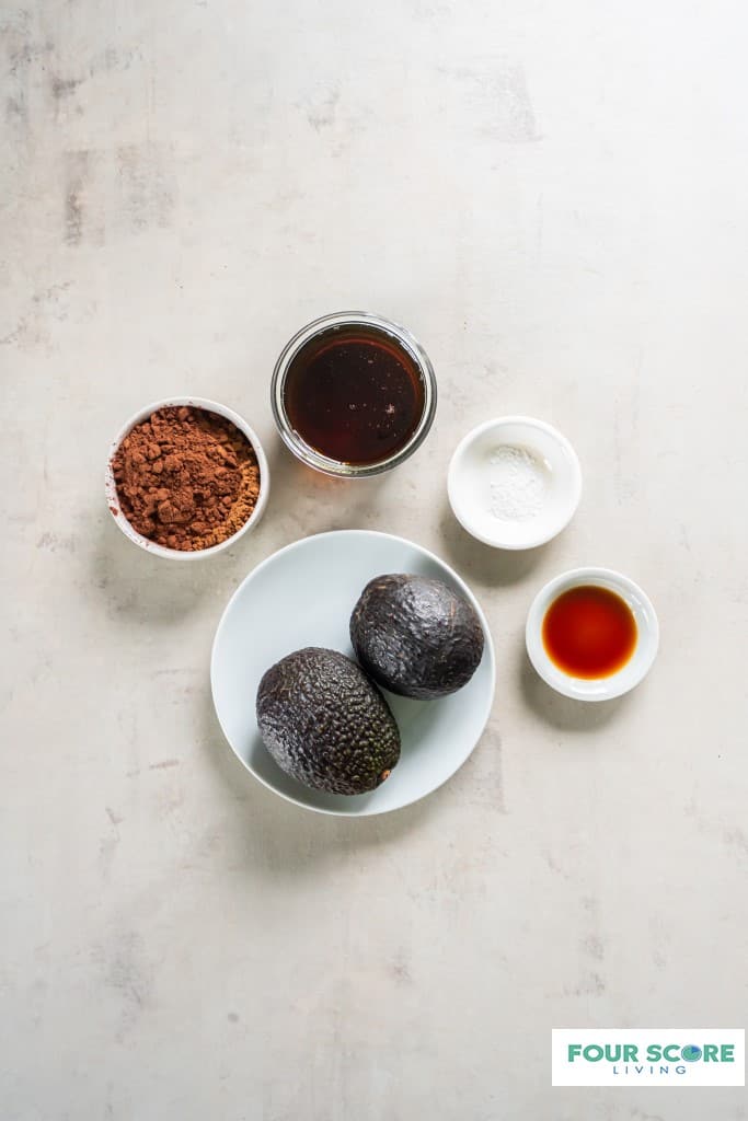 On a white surface there are four small bowls, one with vanilla extract one with salt, one with maple syrup and one with cocoa powder.  A small white dish holding 2 hass avocados is also on the white surface. 