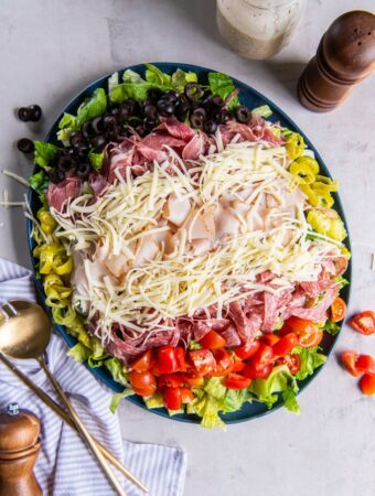 Aerial shot of prepared grinder salad on a plate, all ingredients layered including romaine lettuce, red cherry tomatoes, shredded cheese, sliced turkey, salami and capicola. On the surface there is a wooden seasoning grinder and two brass serving utensils as well as a pin striped cotton kitchen towel.