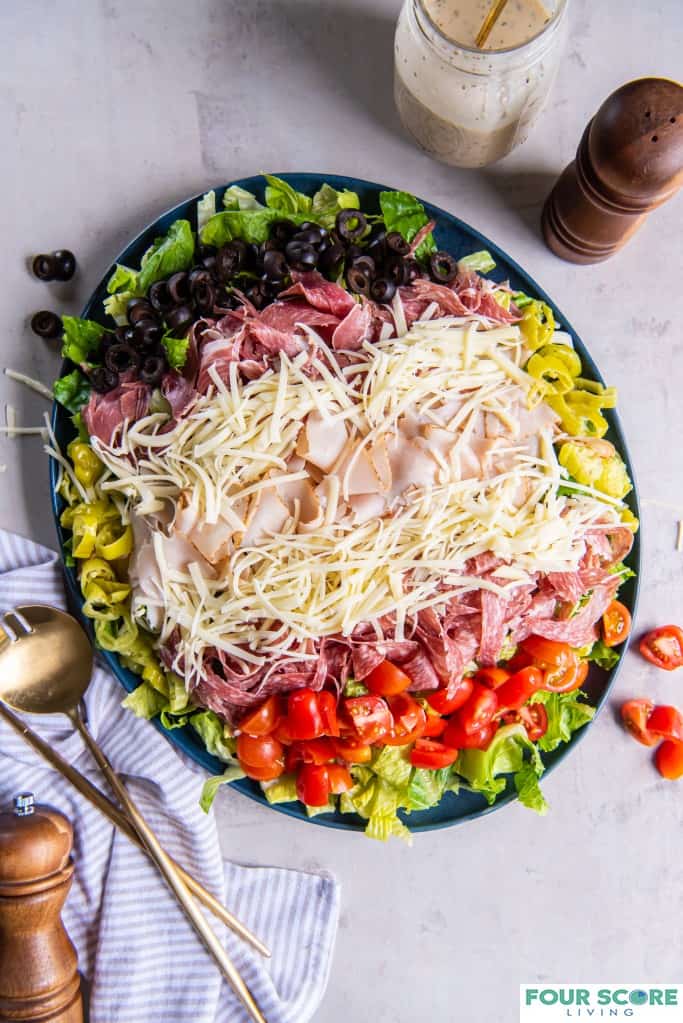 Aerial shot of prepared grinder salad on a plate, all ingredients layered including romaine lettuce, red cherry tomatoes, shredded cheese, sliced turkey, salami and capicola. On the surface there is a wooden seasoning grinder and two brass serving utensils as well as a pin striped cotton kitchen towel. 