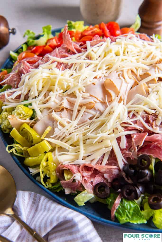 Diagonal shot of prepared grinder salad on a plate, all ingredients layered including romaine lettuce, red cherry tomatoes, shredded cheese, sliced turkey, salami and capicola. On the surface there is a wooden seasoning grinder and two brass serving utensils as well as a pin striped cotton kitchen towel. A glass jar of dressing is also visible in the background.