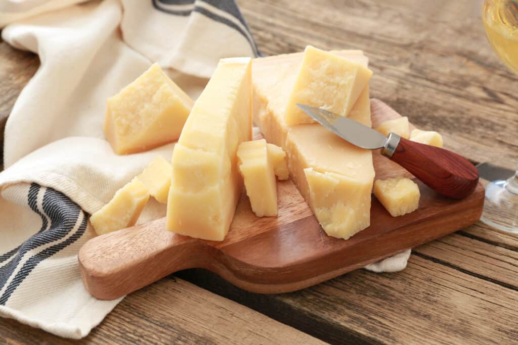 Cutting board with Parmesan cheese on wooden table with a cheese knife and a dish towel.