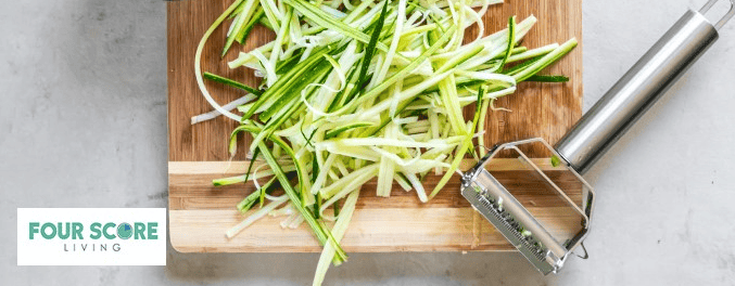 part of a wooden cutting board showing with sliced zucchini noodles on it and a julienne peeler.