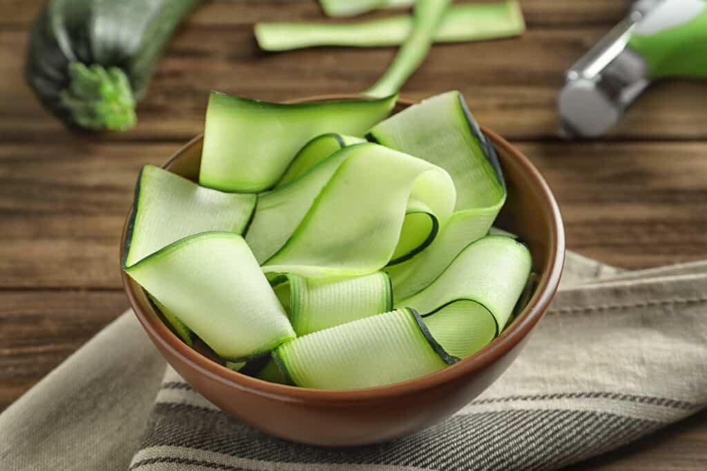 Bowl placed on top of dish towel with wide zucchini noodle slices sitting on a  wood table surface.