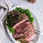 White oval dish with a cooked sirloin tip roast placed on top. Half of the sirloin tip is sliced into slices and a two tong serving tool is piercing the slices. There is a green garnish on the plate, a natural color kitchen towel and wooden salt and pepper shakers in the background.