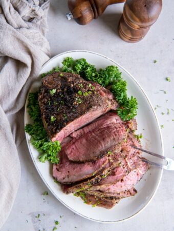 White oval dish with a cooked sirloin tip roast placed on top. Half of the sirloin tip is sliced into slices and a two tong serving tool is piercing the slices. There is a green garnish on the plate, a natural color kitchen towel and wooden salt and pepper shakers in the background.