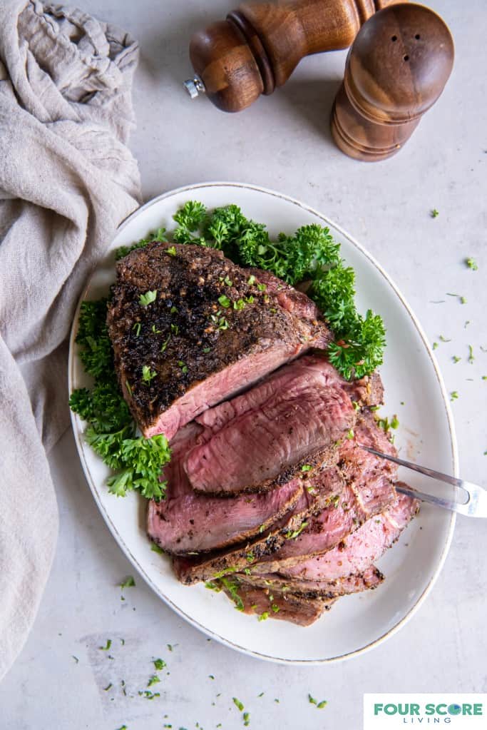 White oval dish with a cooked sirloin tip roast placed on top. Half of the sirloin tip is sliced into slices and a two tong serving tool is piercing the slices. There is a green garnish on the plate, a natural color kitchen towel and wooden salt and pepper shakers in the background.