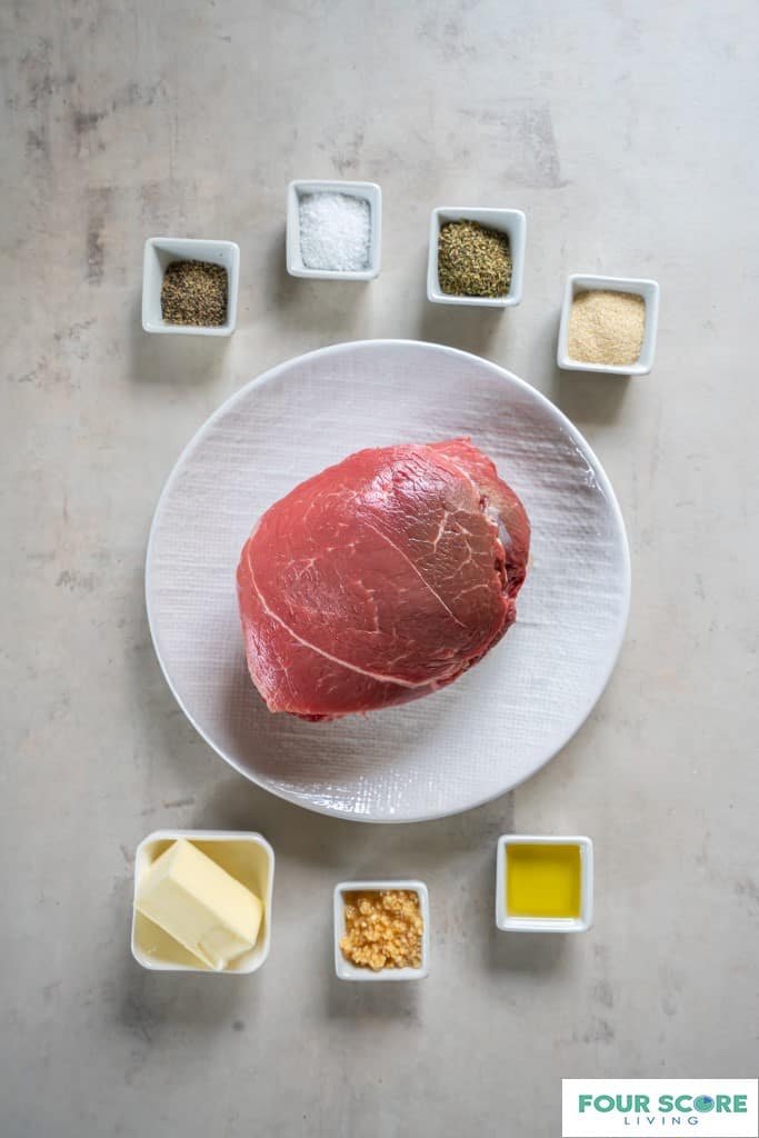 A raw sirloin tip roast placed on a round white plate. On the surrounding surface there are seven small, square white dishes with additional ingredients: butter, oil, minced garlic, salt, spices. 