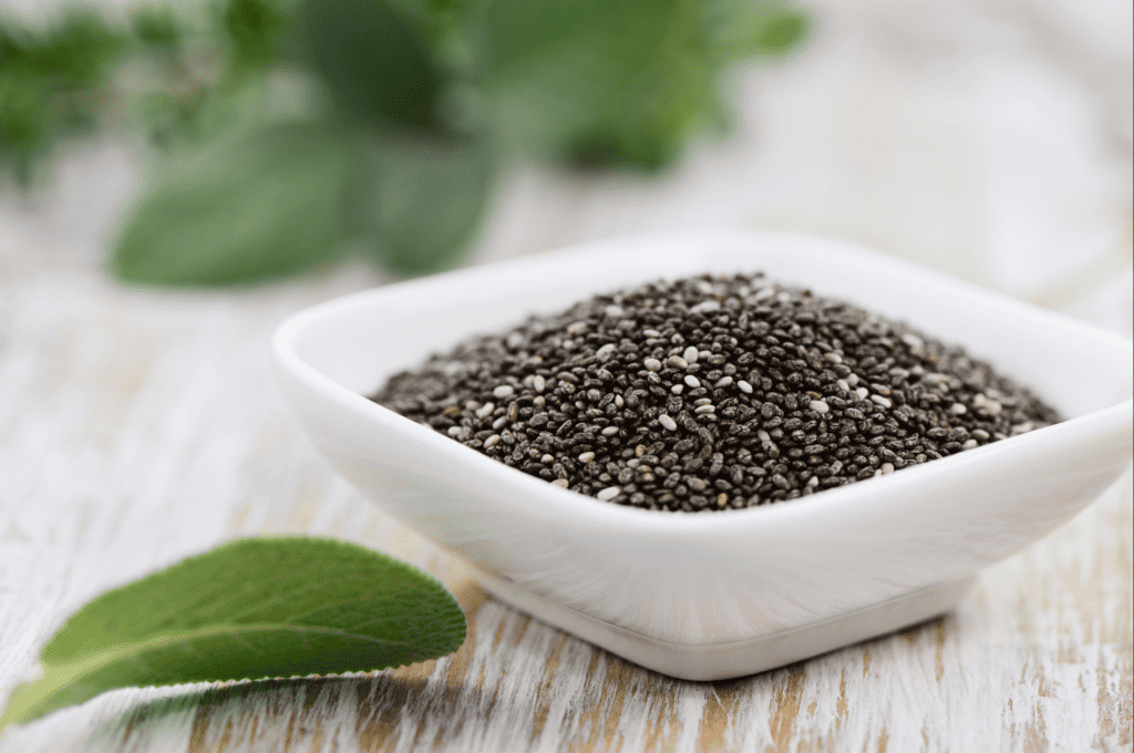Square white dish of chia seeds on a wooden surface with green leaf garnish on the surface