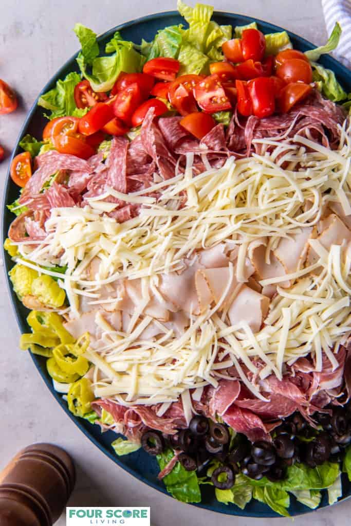 Diagonal shot of prepared grinder salad on a plate, all ingredients layered including romaine lettuce, red cherry tomatoes, shredded cheese, sliced turkey, salami and capicola. On the surface there is a wooden seasoning grinder and two brass serving utensils as well as a pin striped cotton kitchen towel. A glass jar of dressing is also visible in the background.