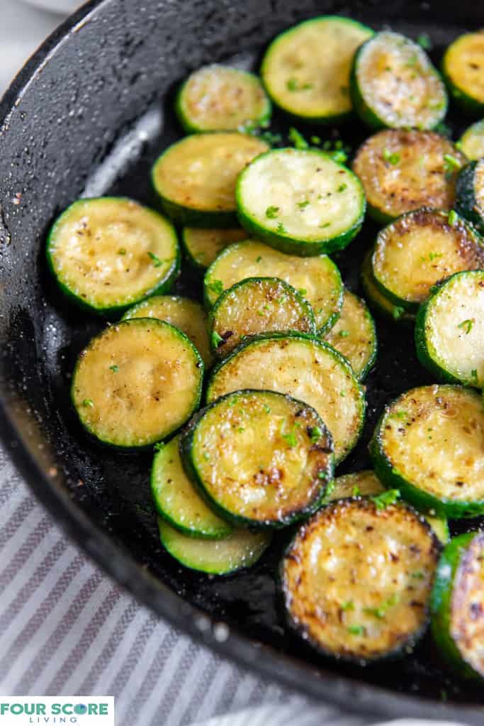 Close up of sauteed zucchini rounds in a cast iron pan with a pinstriped kitchen towel in the background.