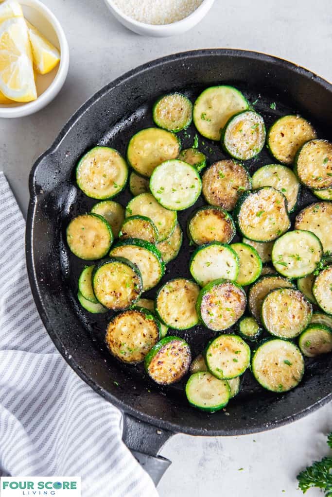 Rounds of sauteed zucchini in a cast iron pan eith a white dish of lemon wedges, a pinstriped kitchen towel, green garnish, and a white dish of grated parmesan cheese