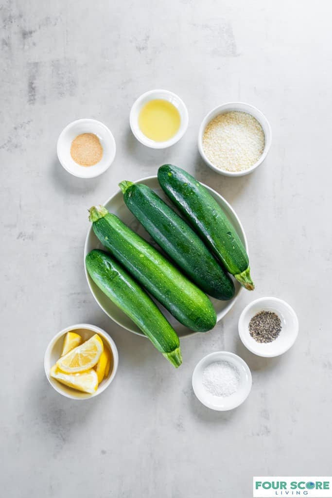 Medium sized white bowl with four whole medium zucchini across the bowl. Six small white bowls with seasonings, oil, parmesan cheese, and lemon wedges.