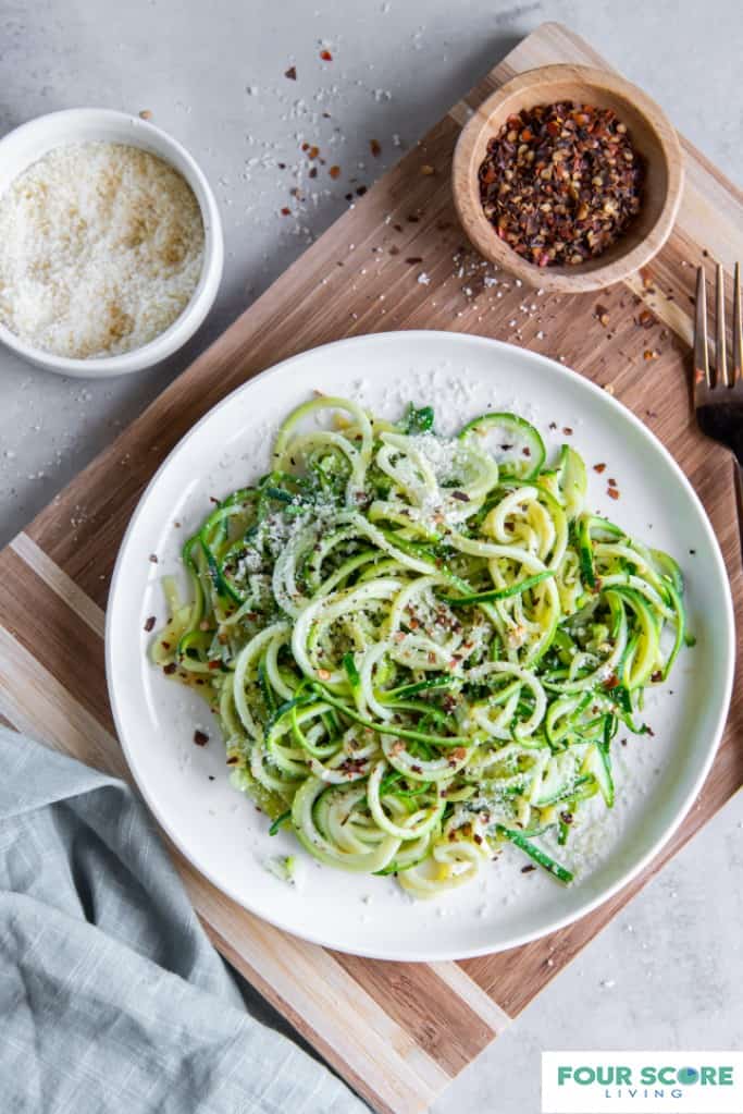 Wooden cutting board with white plate sitting on top. Cooked zucchini noodles are on the white plate sprinkled with parmesan cheese. There is a light grey dish towel and a brass fork nearby. There is a small wooden bowl of red pepper flakes and a small white bowl of grated parmesan cheese.