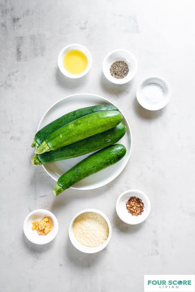 White plate with 4 whole fresh zucchini on top. Six small white bowls each with an ingredient. Red pepper flakes, minced fresh garlic, parmesan cheese, salt, Black pepper, avocado oil. 